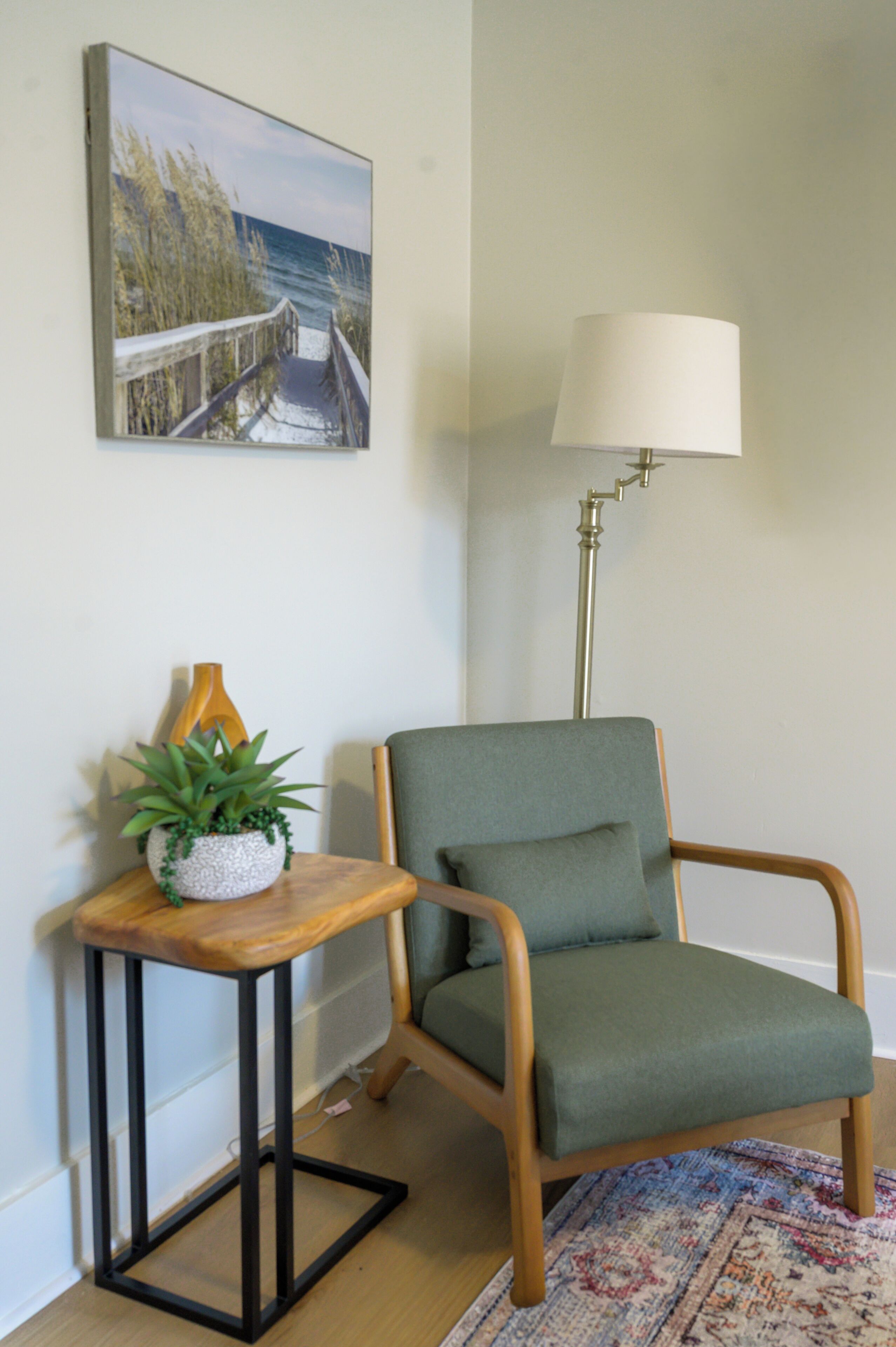 A room with a wooden chair, a small table with a plant, a lamp, and a painting on the wall.