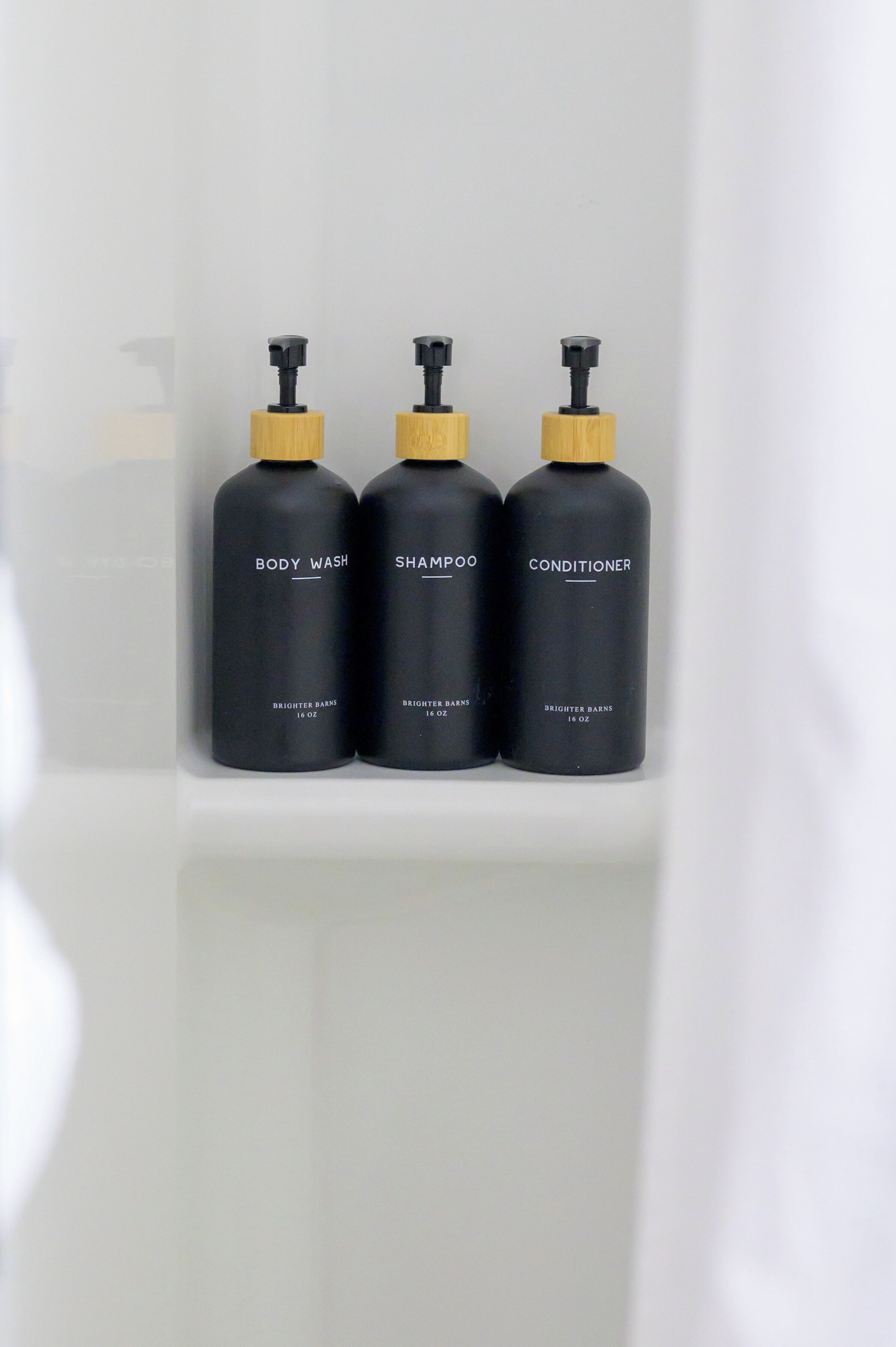 Three black bottles with yellow caps labeled 'BODY WASH,' 'SHAMPOO,' and 'CONDITIONER' on a white shelf.