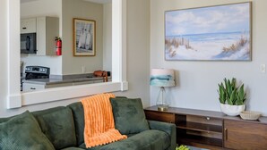 Una sala de estar con un sofá verde, una mesa redonda de madera, un cuadro de una playa y una cocina visible a través de un arco.