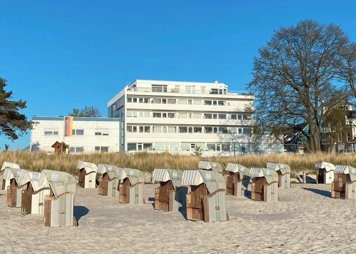 A row of beach huts numbered 37 to 69 on a sandy beach, with a large building in the background.