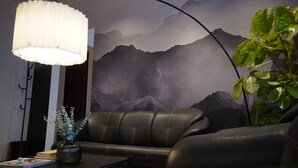 A modern living room with a black leather sofa, a table with a vase of flowers, a wall-mounted lamp, and a large wall mural of mountains.
