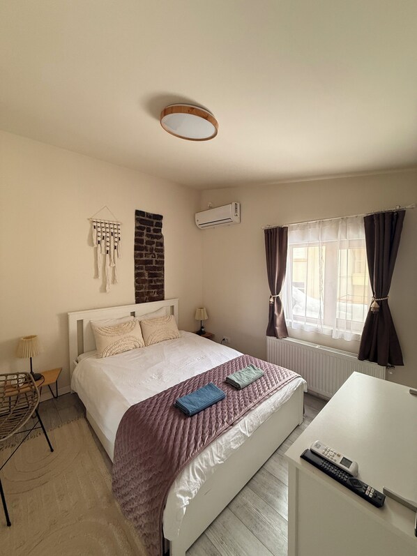 Un dormitorio con cama, mesita de noche, lámpara, aire acondicionado y ventana con cortinas.