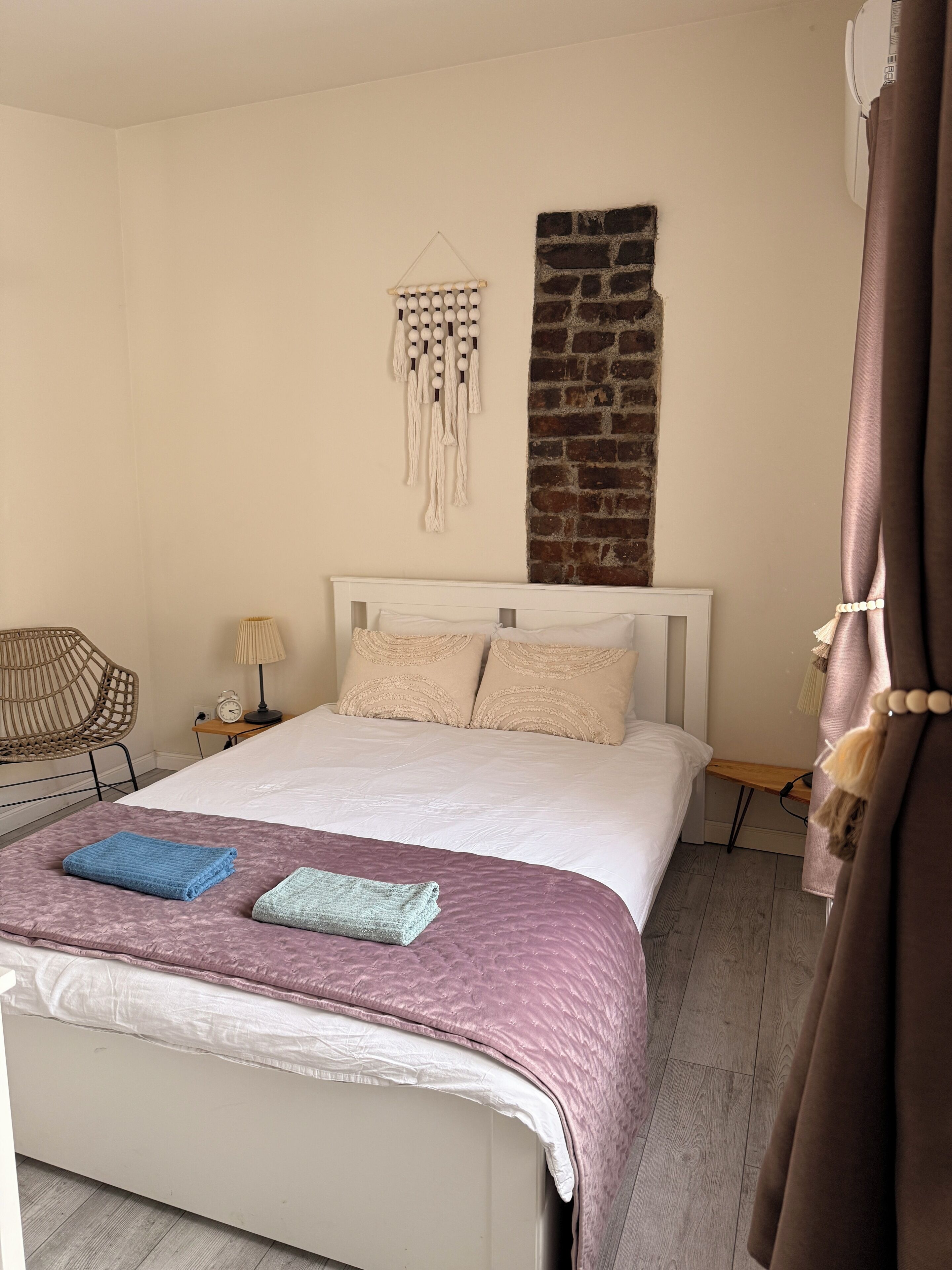 Un lit bien fait, recouvert d’une couverture rose, une décoration murale en macramé et un mur d’accent en briques.