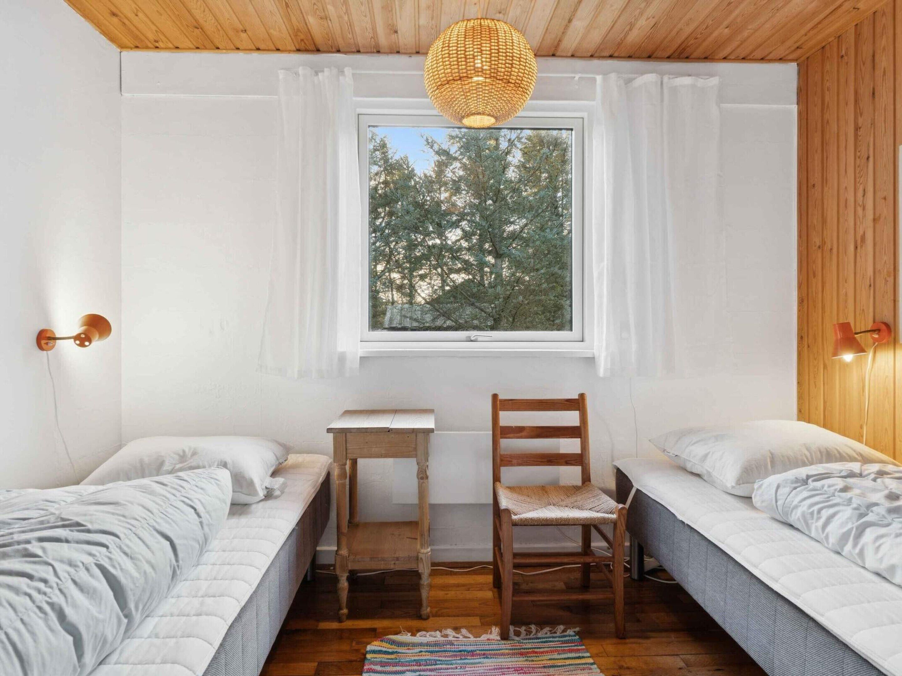 A room with two beds, a small wooden table, a chair, a window with a view of trees, and a woven light fixture.