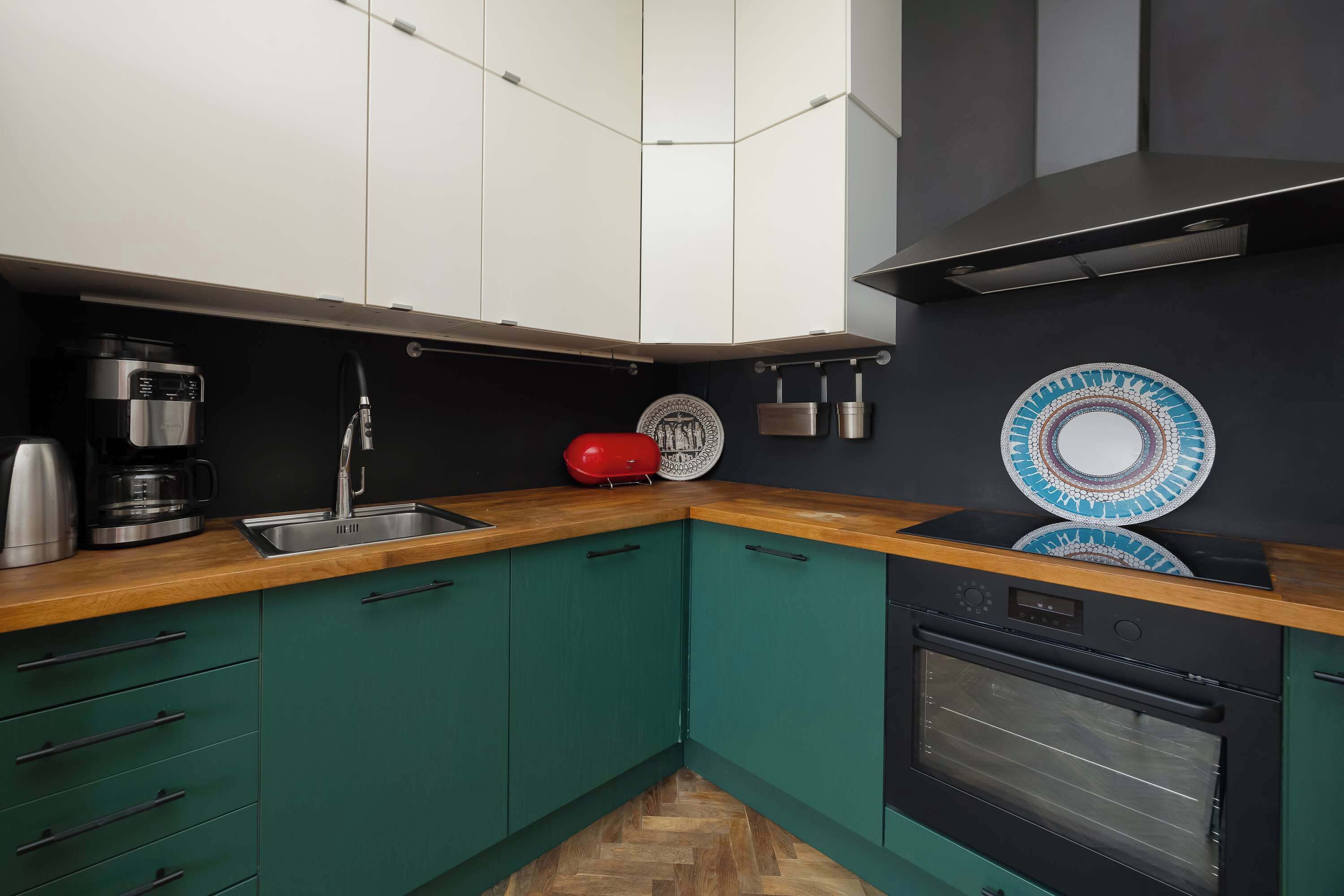 Una cocina con armarios verde azulado, encimera de madera, cafetera, hervidor rojo y horno empotrado.