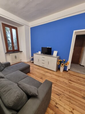 Ein Wohnzimmer mit grauem Sofa, einem Fernseher auf einer weißen Kommode und einer blauen Wand.
