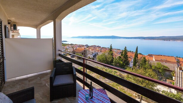 Un balcon agrémenté de meubles en osier, une table avec une bouteille et des verres, et une vue sur une ville côtière et la mer.