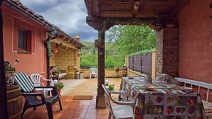 An outdoor patio with a table covered in a vegetable print tablecloth, surrounded by white plastic chairs and a wooden bench.
