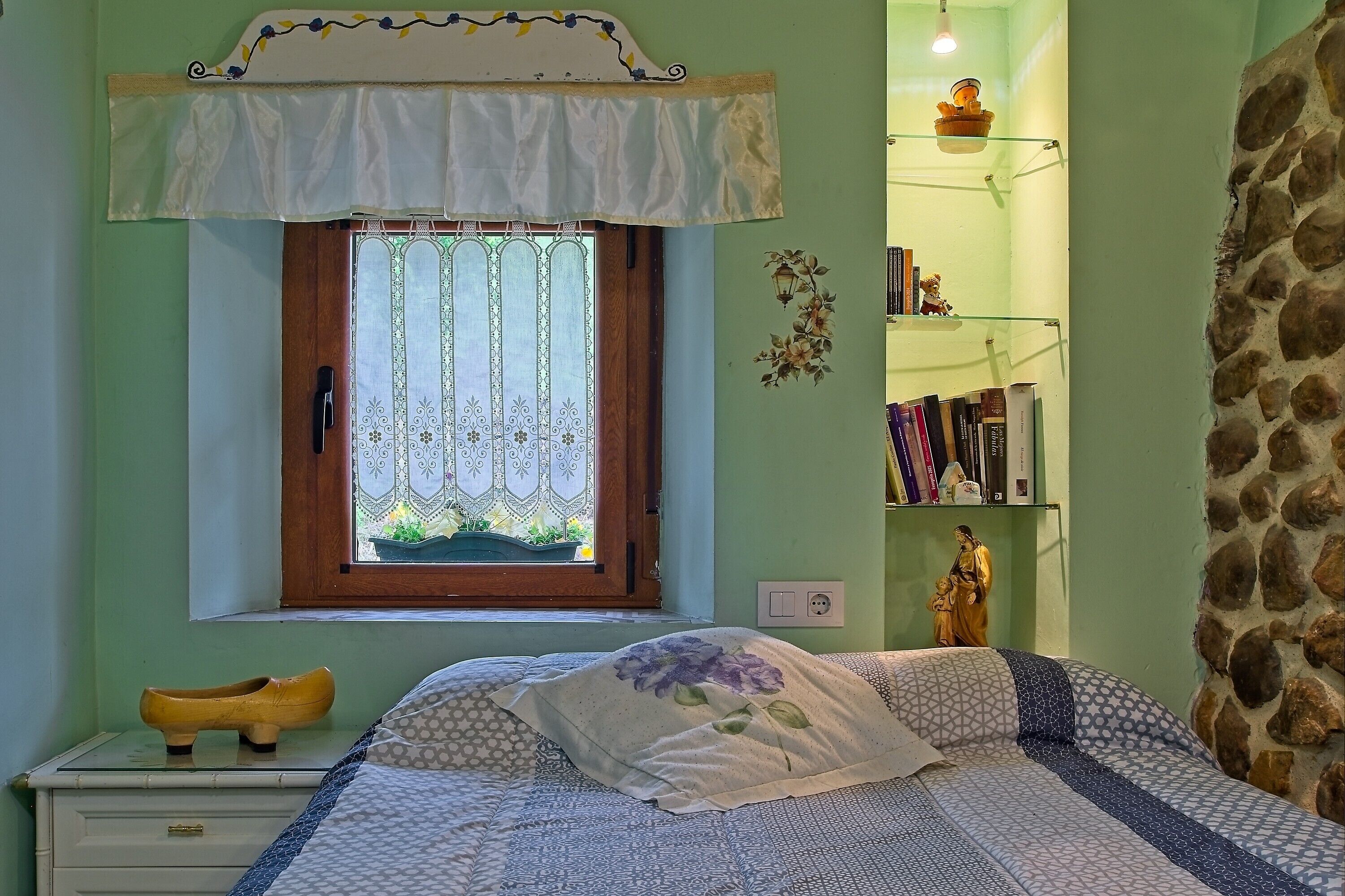 A bedroom with a bed, a window with curtains, a nightstand with a shoe, a statue, and a bookshelf.