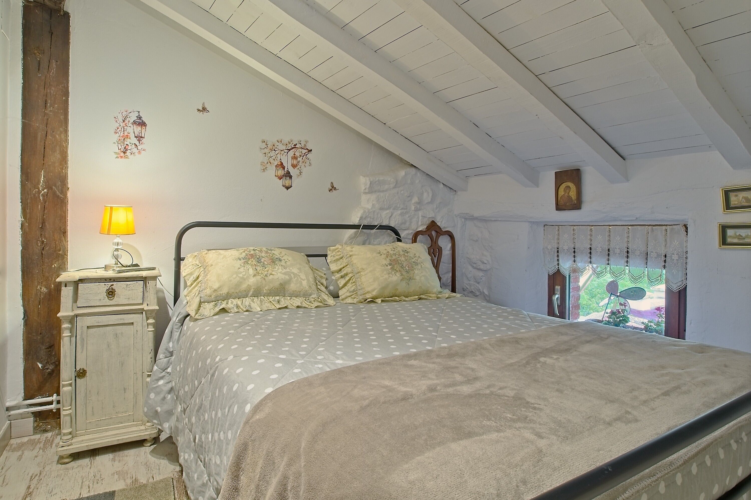 A bedroom with a bed, a nightstand, a lamp, a window with curtains, and a wall with decorative elements.