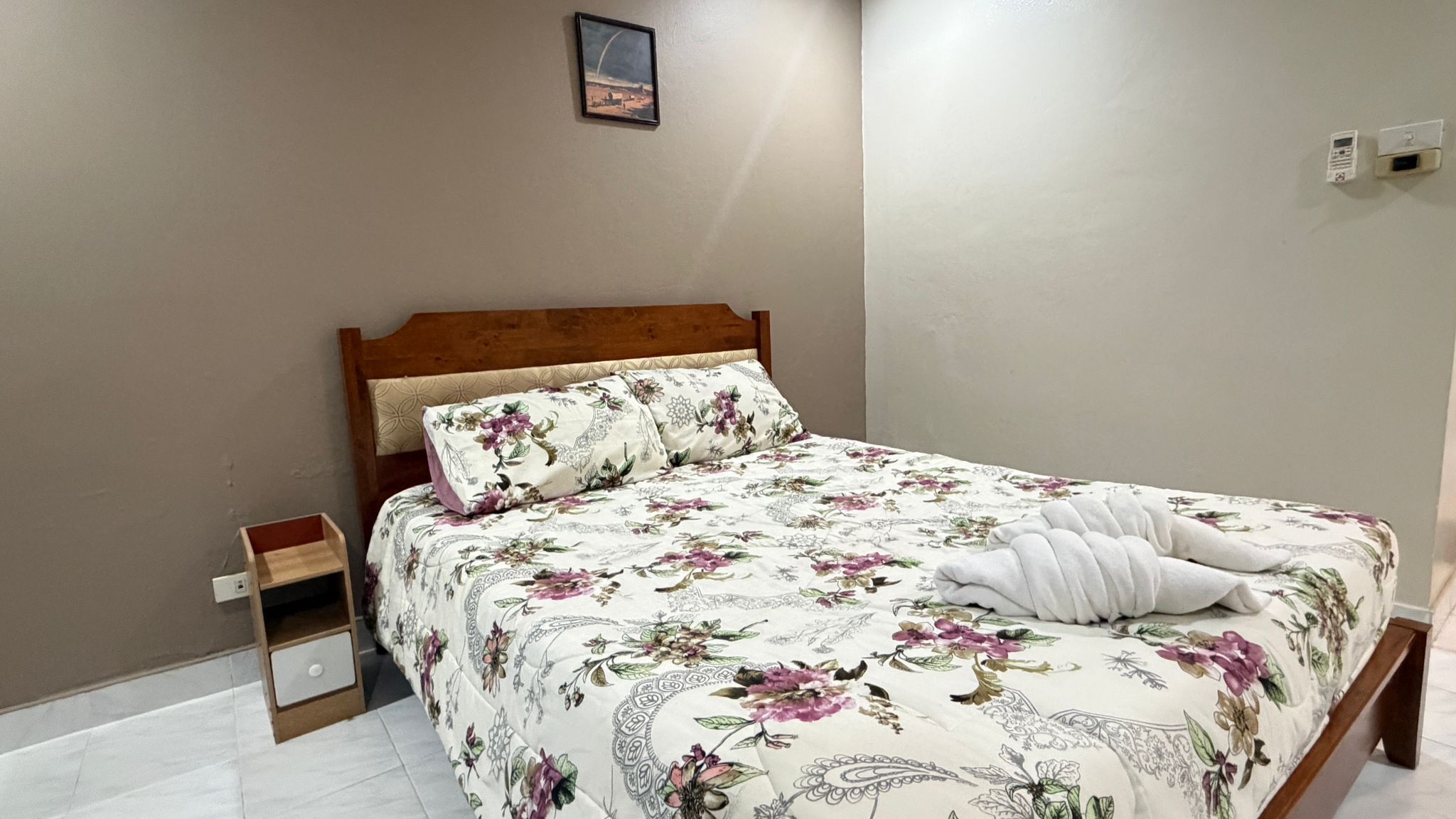 A neatly made bed with floral bedding, a wooden headboard, a small bedside table, and a wall-mounted picture.