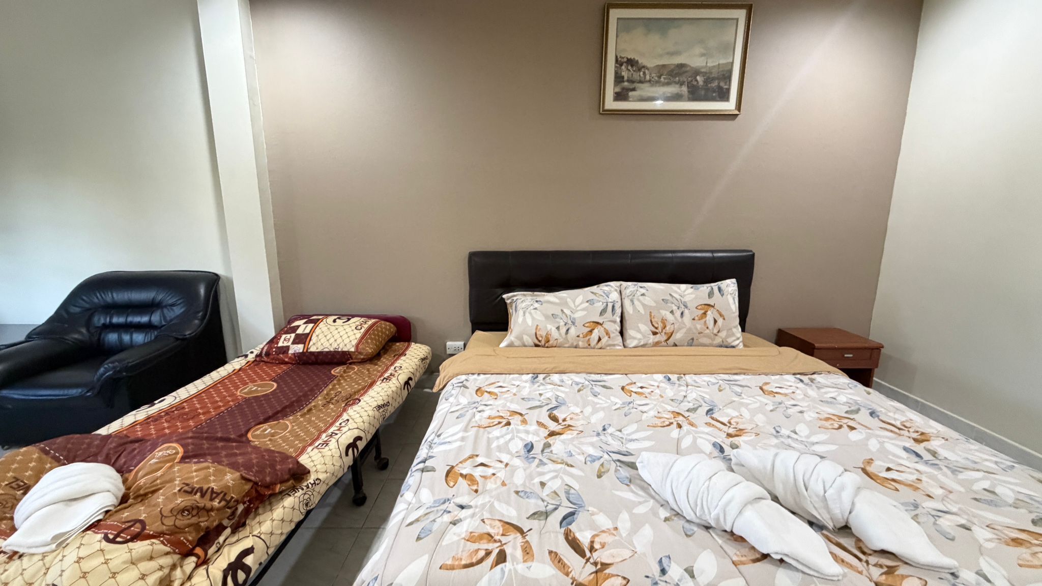 A hotel room with two single beds, a black leather chair, a small wooden nightstand, and a framed picture on the wall.