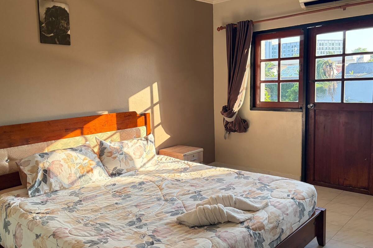 Un dormitorio con una cama de madera, ropa de cama floral, una mesita de noche, una puerta de madera y una ventana con cortinas.