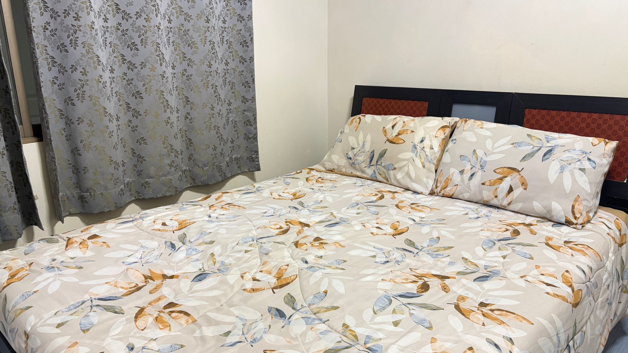 A bed with a floral bedspread, a headboard with a patterned panel, and a window with curtains.