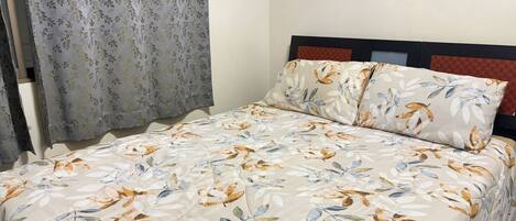 A bed with a floral bedspread, a headboard with a patterned panel, and a window with curtains.