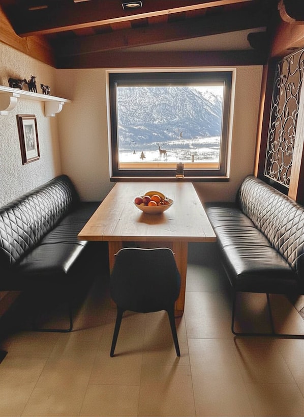 A dining area with a wooden table, two black leather booths, a bowl of fruit, and a window with a mountain view. - Landhaus Osttirol | Apartement Balkony (Göriach)