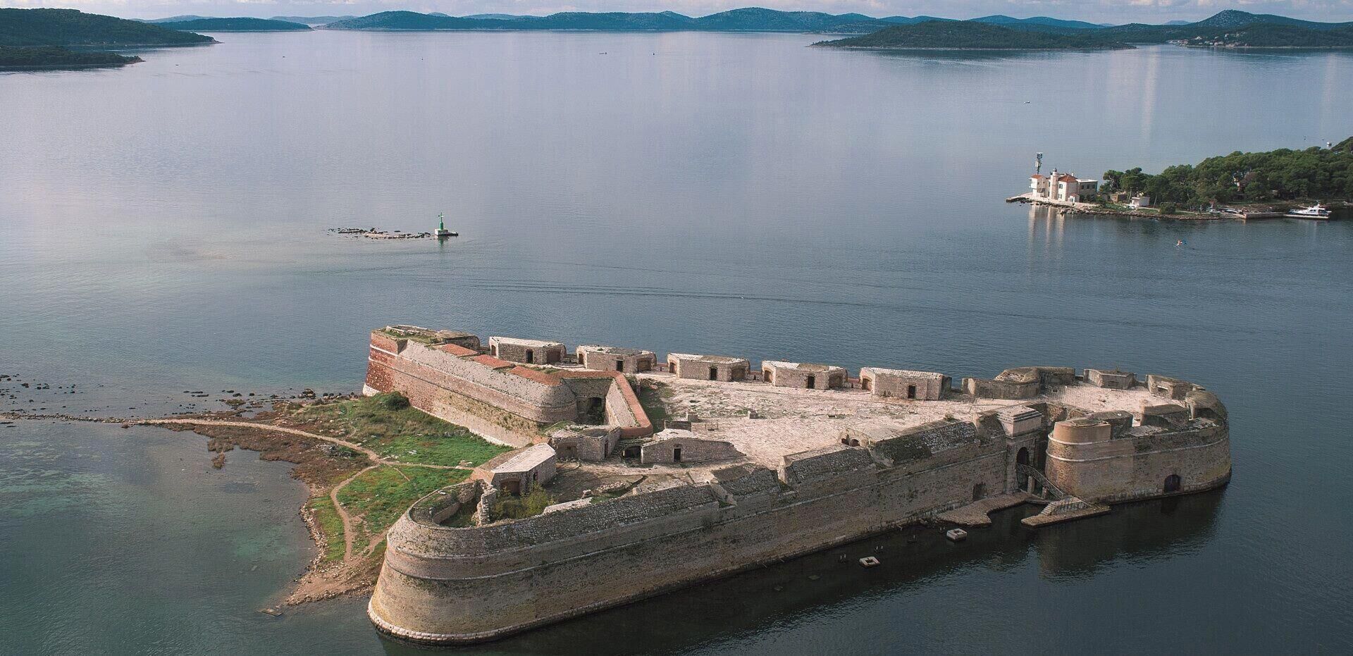 Vue aérienne d’une forteresse historique dotée d’un fossé, entourée d’eaux calmes et de collines lointaines.