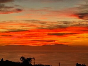 Un atardecer sobre un paisaje costero con siluetas de palmeras y montañas a lo lejos.