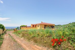 A house with a red roof, a dirt path leading to it, and a view of the sea in the background.