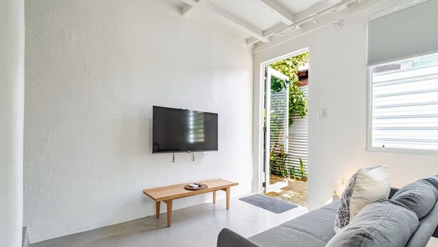 Uma sala de estar moderna com uma TV de tela plana, um banco de madeira, um sofá cinza e uma porta de vidro deslizante que dá para um jardim.