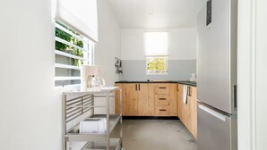 Cozinha estreita com frigorífico embutido, armários de madeira, máquina de café e janela com vista para vegetação.