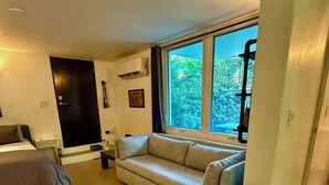 Un salón moderno con un sofá, un sillón azul, una cama y un amplio ventanal con vistas a árboles.