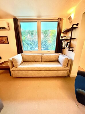 Una sala de estar con un sofá, una ventana con cortinas, un estante de pared con libros y un cuadro enmarcado.