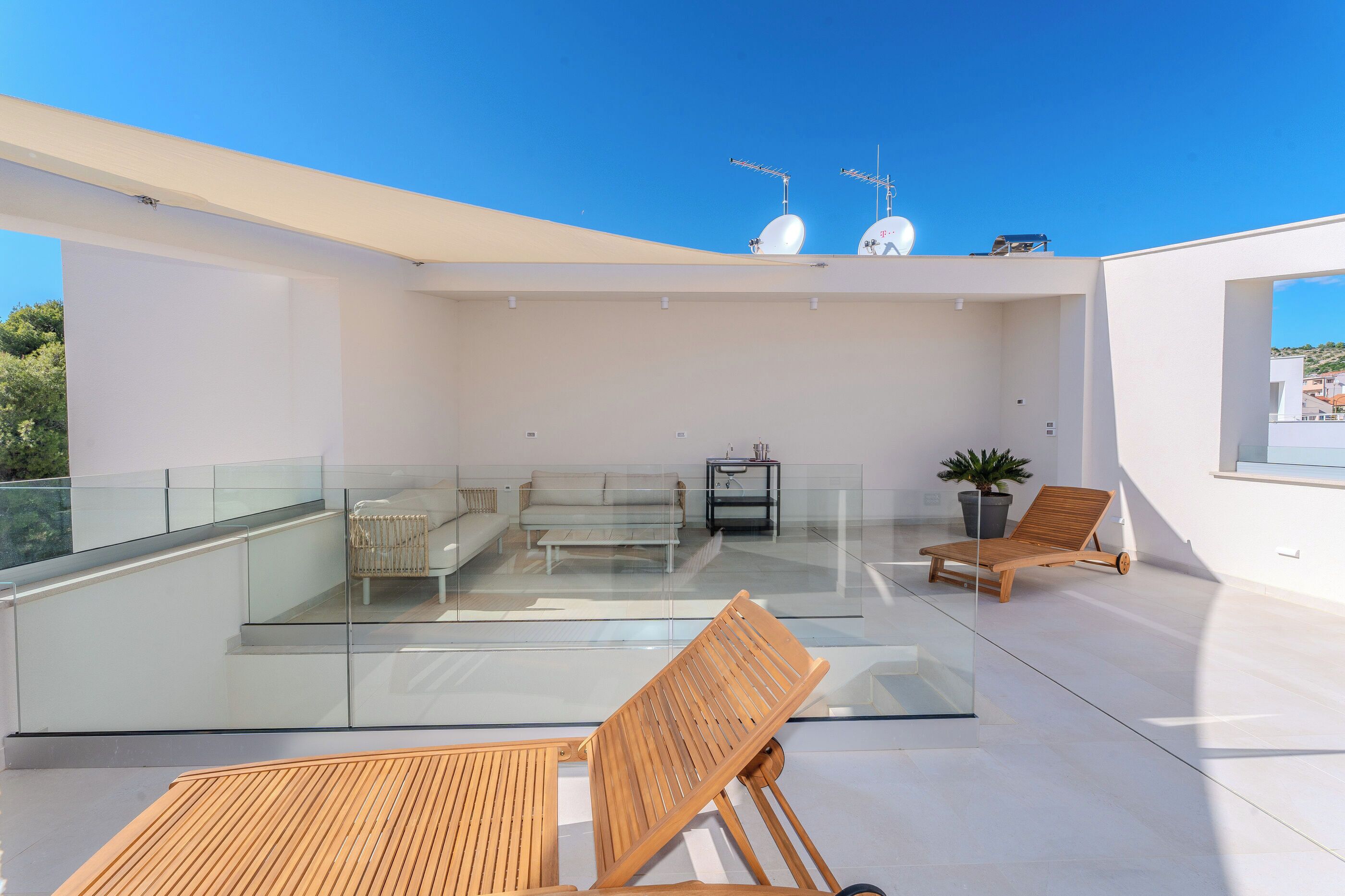 A modern rooftop terrace with glass railings, a sofa, a wooden chair, and a potted plant.