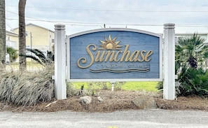 Schild für die Sunchase Condominiums mit Sonnensymbol, umgeben von Bepflanzung und Wohngebäuden.