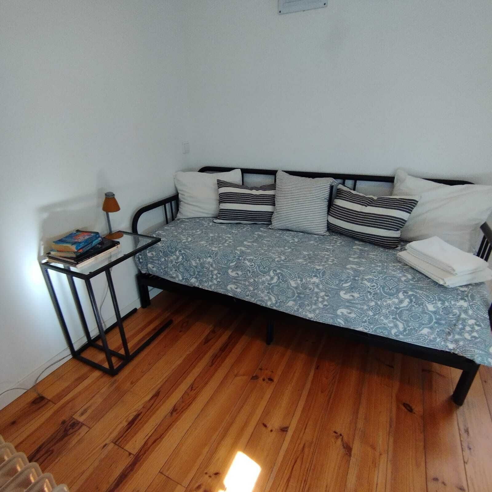 A single bed with a patterned bedspread, a bedside table with books, and a lamp.