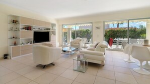 Una sala de estar moderna con televisor de pantalla plana, sofás blancos, una mesa de centro de vidrio y un balcón con mobiliario exterior.
