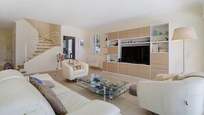 Una sala de estar moderna con una mesa de centro de vidrio, sofás blancos, una escalera y estanterías empotradas.