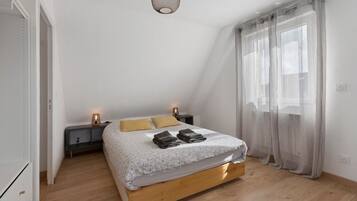 A bedroom with a bed, two bedside tables, a lamp, a window with curtains, and a light fixture.