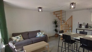 A living room with a sofa, a coffee table, and a dining area with a table and chairs. There is a staircase leading to the upper floor. - 2-Star Holiday Home “Le Mascaret des Abbayes du Val de Seine” (Sainte-Marguerite-sur-Duclair)