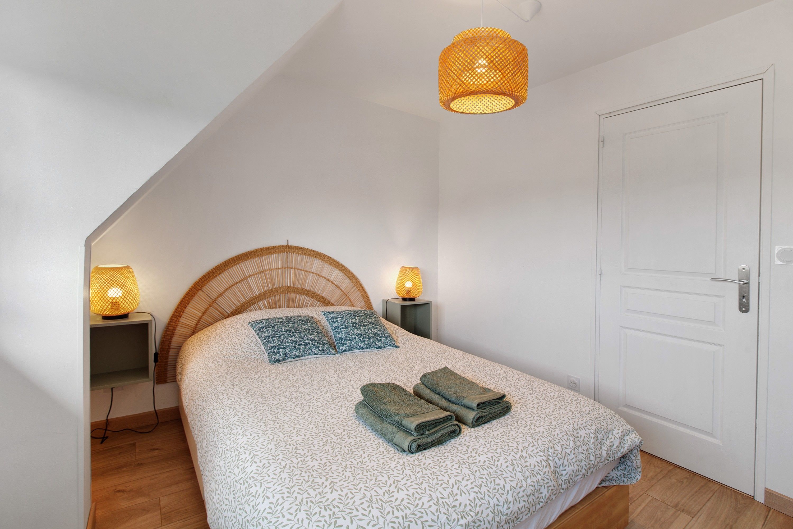 Une chambre avec un plafond en pente, un lit avec une literie à motifs, deux lampes de chevet et une porte.