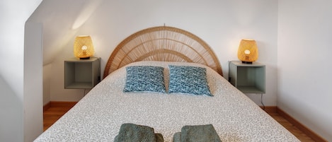A bedroom with a sloped ceiling, a bed with patterned bedding, two bedside lamps, and a woven headboard.