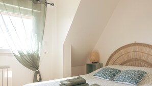 A bedroom with a bed, a window with a sheer curtain, a bedside lamp, and a rattan headboard. - 2-Star Holiday Home “Le Mascaret des Abbayes du Val de Seine” (Sainte-Marguerite-sur-Duclair)