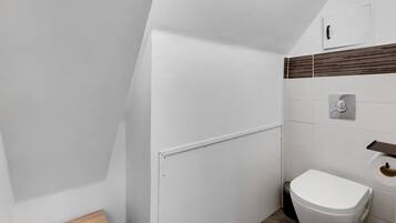 Bany petit i modern amb vàter blanc, una cadira de fusta i paret amb rajoles i un panell decoratiu de fusta.