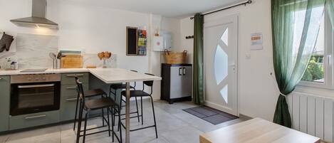 A modern kitchen with a built-in oven, a dining area with bar stools, and a view through a window with green curtains.