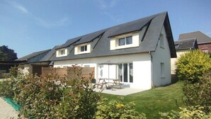 Une maison à deux étages avec un toit en pente, une terrasse avec une table et des chaises, et un jardin agrémenté d’arbustes et d’un petit arbre.