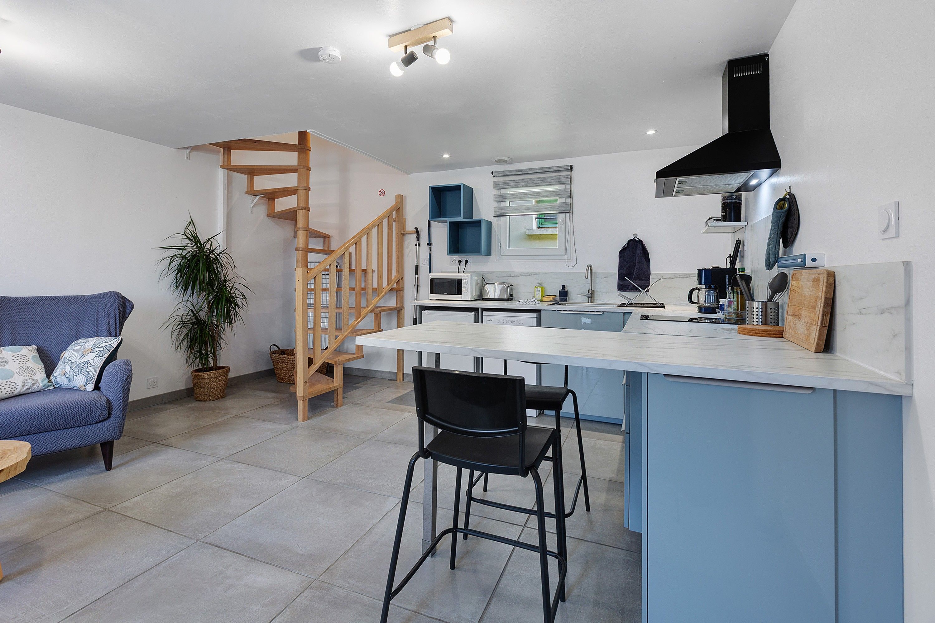 A modern kitchen with a central island, wooden staircase, and a blue sofa.