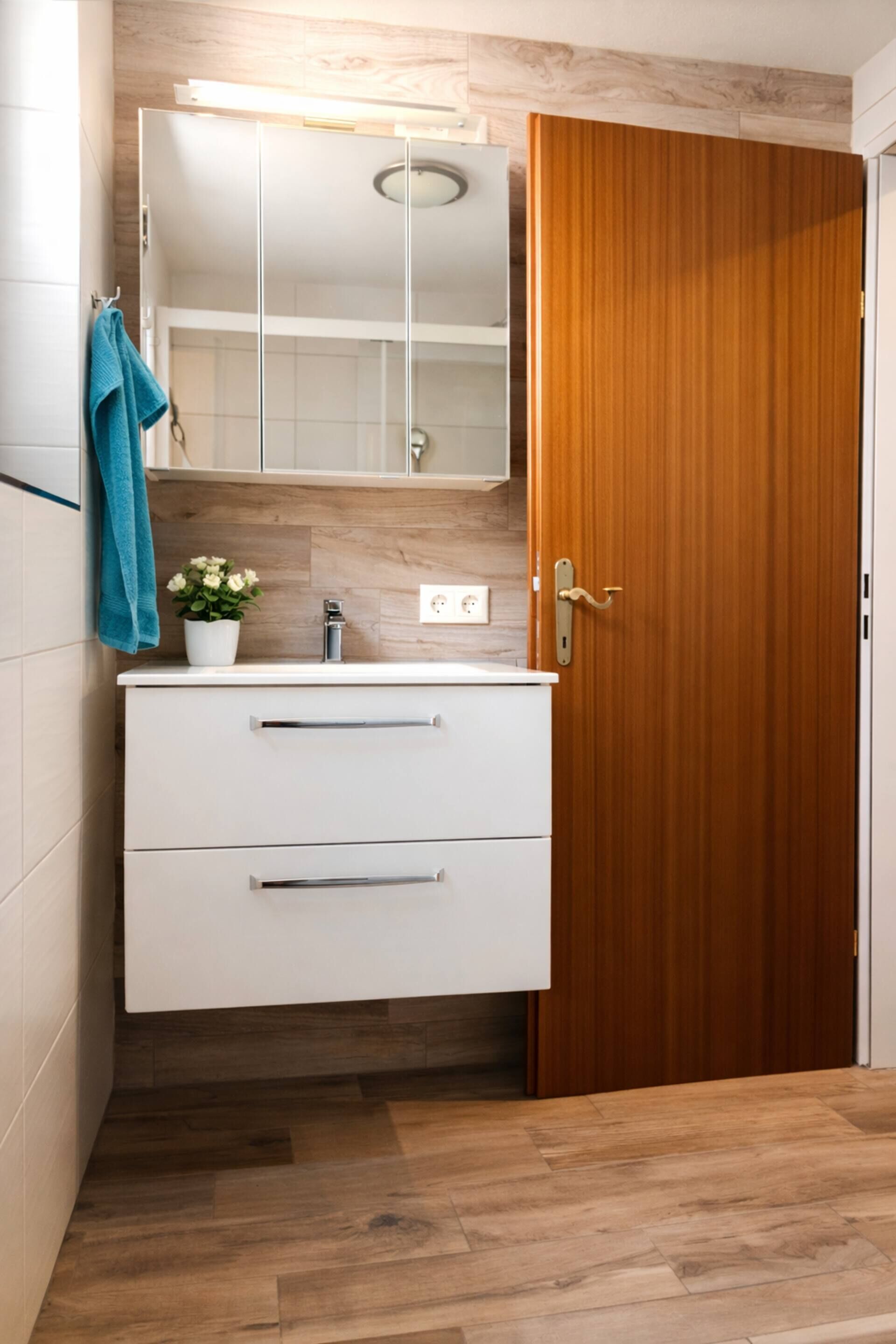 Ein Badezimmer mit Holztür, einem weißen Waschtisch mit Schubladen, einem Spiegel über dem Waschtisch und einer Topfpflanze auf dem Waschtisch.
