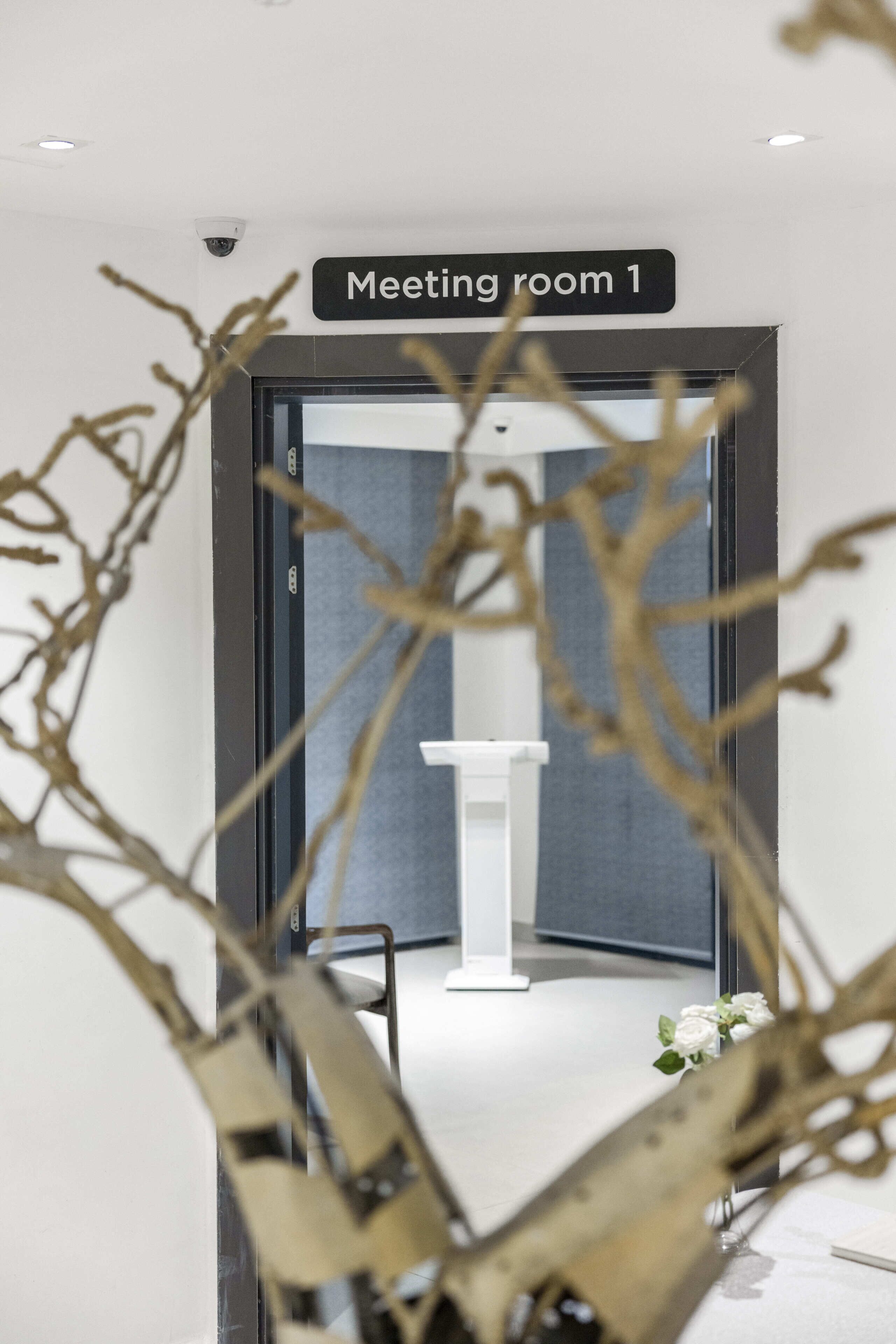 A doorway to a meeting room with a sign above it, a white pedestal, and a vase with white flowers.