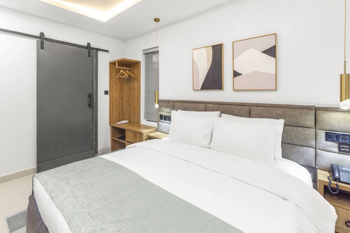 A modern hotel room with a large bed, a wardrobe, a bedside table with a phone, and abstract wall art.