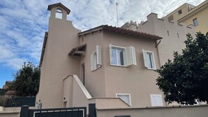 Un edificio con fachada color beige, contraventanas blancas, un pequeño balcón y una entrada protegida por un portón.