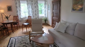 Ein Wohnzimmer mit einer Couch, einem Esstisch mit Stühlen, einem Dekorschaukelstuhl, einem kleinen Tisch mit einer Vase und einem Fenster mit Vorhängen.