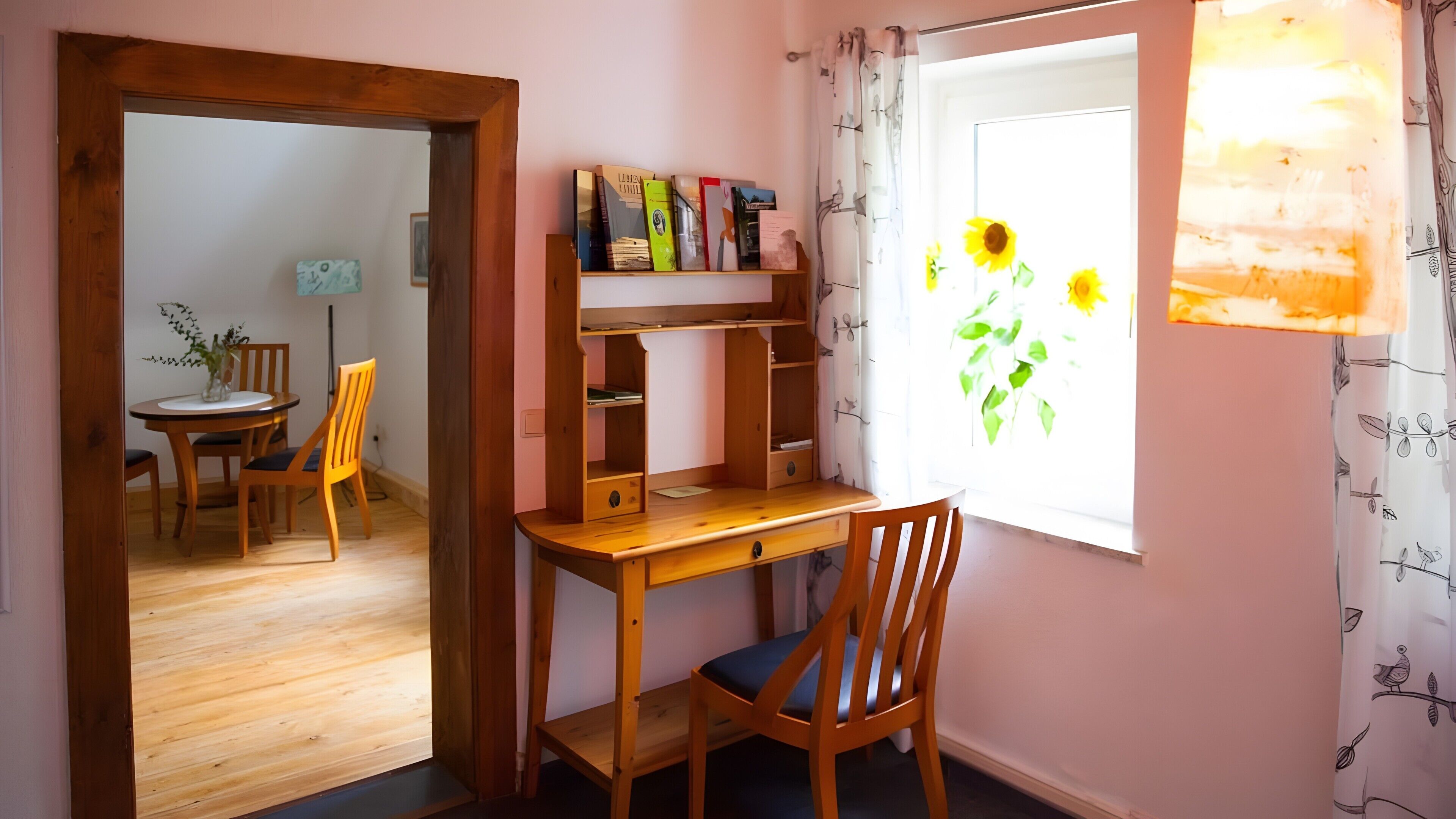 Una habitación con un escritorio de madera, una silla, una estantería, un comedor con sillas y una mesa, y una ventana con girasoles.