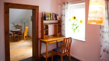 Una habitación con un escritorio de madera, una silla, una estantería, un comedor con sillas y una mesa, y una ventana con girasoles.