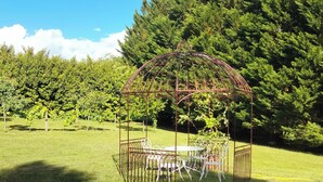 Um gazebo de metal com uma mesa e cadeiras em uma área gramada, com árvores ao fundo.