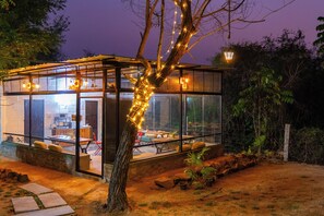 Une structure entièrement vitrée comprenant un espace repas, des sièges en plein air et un arbre décoré de guirlandes lumineuses.